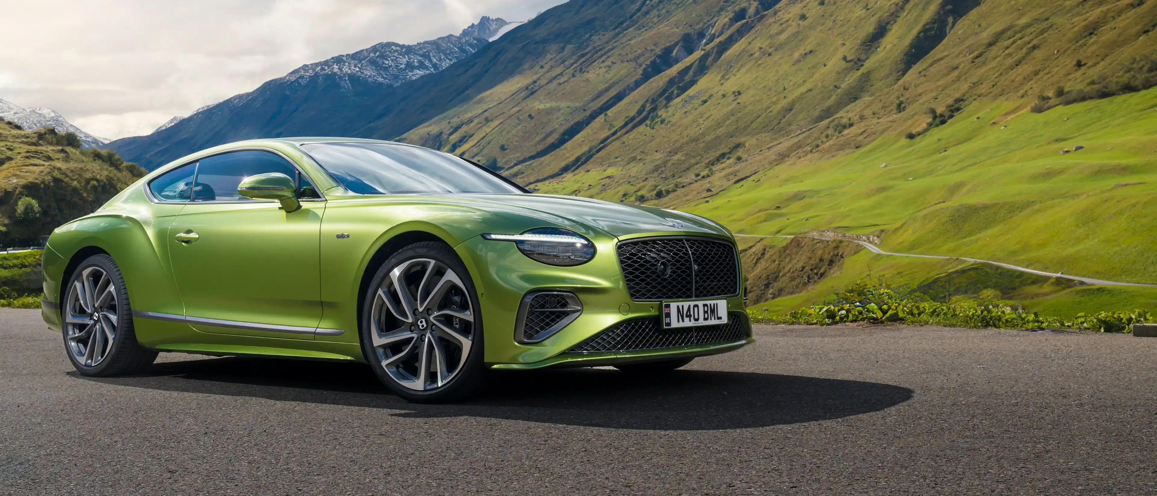Side profile of a lime-green Bentley Continental GT parked on a mountain road with sweeping green valleys and peaks.